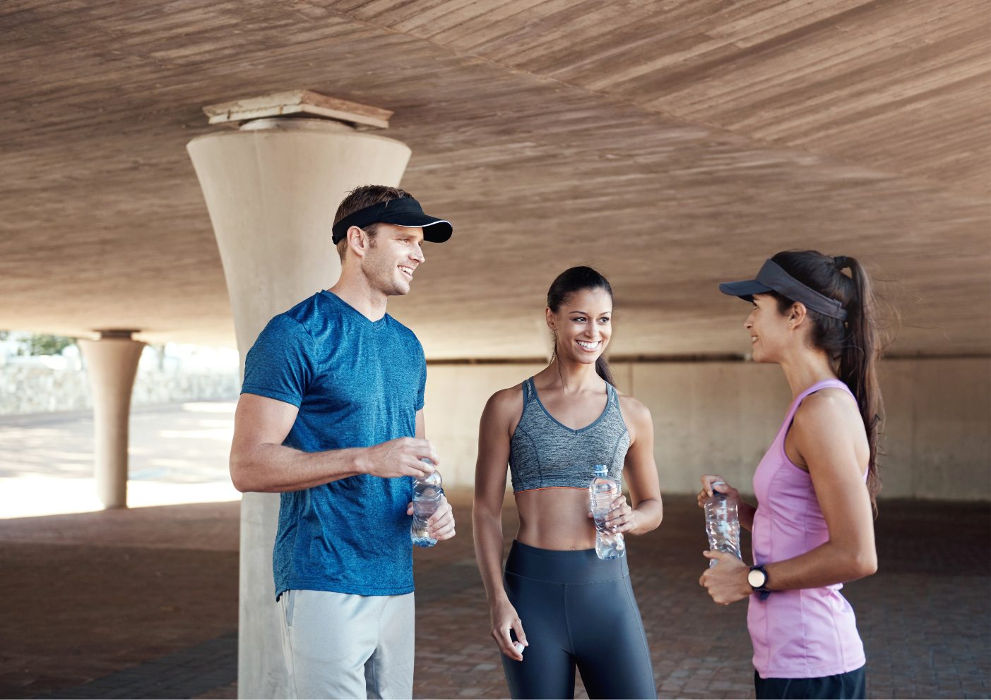 2 vrouwen en 1 man in sportkleding staan onder een tunnel, hebben een flesje water in de hand en 2 hebben er een pet op.