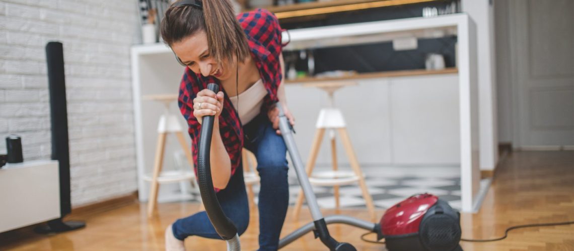 Jonge vrouw is dansend haar huis aan het stofzuigen, gebruikt stofzuigerslang als microfoon en zingt mee