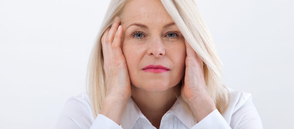 40 plus vrouw in witte blouse kijkt serieus in de camera. Heeft haar handen onder haar hoofd, over haar oren