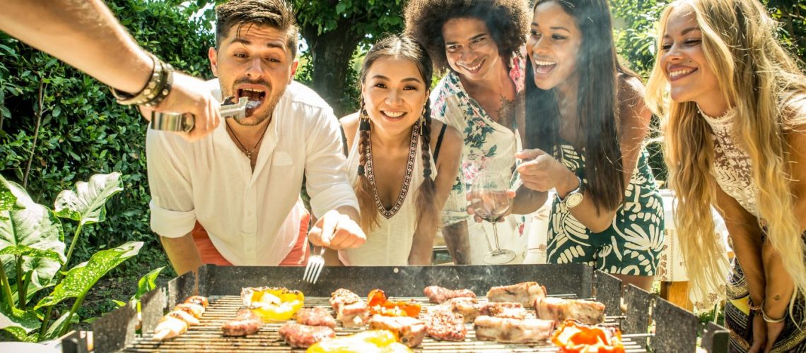 5 vrienden staan bij een BBQ, wachten lachend op een stukje vlees. Een man krijgt alvast een hapje. Zo is er een mannenarm te zien die een vrok vasthoudt met een stuk eten eraan.