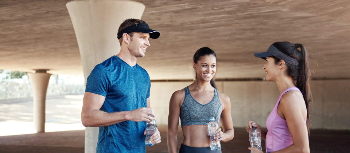 2 vrouwen en 1 man in sportkleding staan onder een tunnel, hebben een flesje water in de hand en 2 hebben er een pet op.